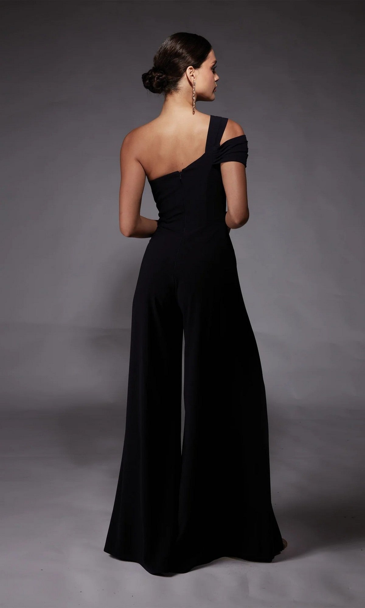 Woman stands with back to camera in the Alyce 70066 Long Formal Jumpsuit, showcasing a chic one-shoulder design and wide-leg pants against a plain gray background.